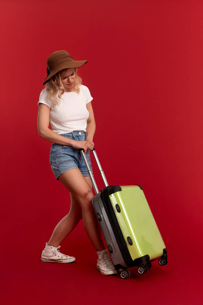 Young blond curly woman in a sundown hat feels tired holding heavy grey luggage bag at sightseeing tour while standing infront of a red background. - Foto, imagen