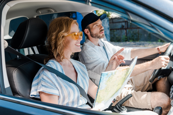 enfoque selectivo de la mujer en gafas de sol apuntando con el dedo mientras sostiene el mapa en el coche cerca del hombre
  - Foto, Imagen