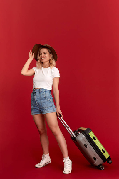 Young blond curly woman in a sundown hat with grey luggage bag is excited at sightseeing tour while standing infront of a red background.  - Fotó, kép