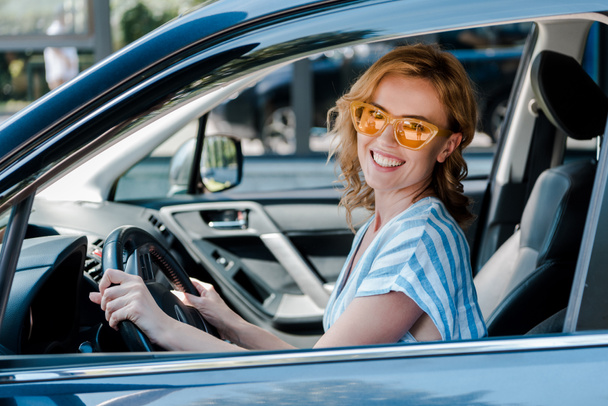 χαρούμενη ξανθιά γυναίκα στα γυαλιά ηλίου κρατώντας τιμόνι, ενώ κάθεται στο αυτοκίνητο  - Φωτογραφία, εικόνα