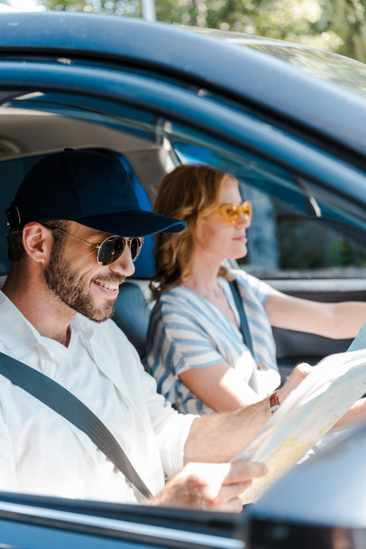 selective focus of happy man in sunglasses and cap holding map near woman in car  - Valokuva, kuva