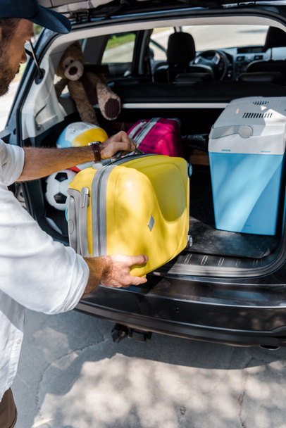 cropped view of man putting yellow luggage near pink travel bag in car trunk  - Фото, зображення