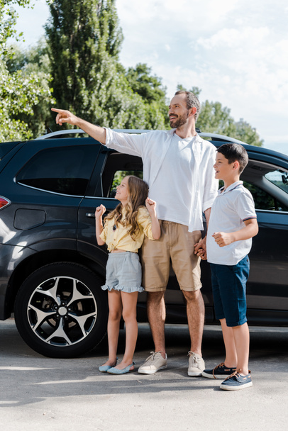 happy father pointing with finger while standing with cheerful kids and car  - Fotoğraf, Görsel