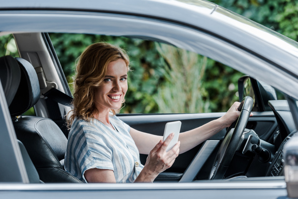 selektivní zaměření usměvavé ženy používající smartphone při jízdě autem  - Fotografie, Obrázek