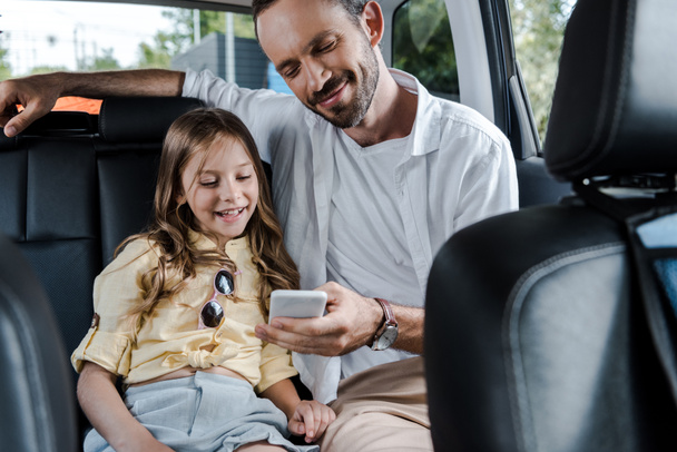 selective focus of happy man using smartphone near daughter in car  - Fotografie, Obrázek