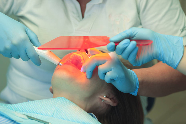 Rellenos dentales fijos y secos del dentista con una luz. Asistente del dentista sosteniendo un vaso naranja para proteger los ojos de un paciente. De cerca.
. - Foto, Imagen