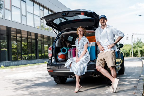 cheerful man in cap standing with hand on hip near woman and car  - Φωτογραφία, εικόνα