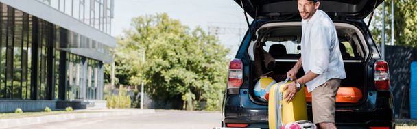 plano panorámico de hombre barbudo alegre en la tapa de pie cerca del equipaje y el coche
  - Foto, Imagen