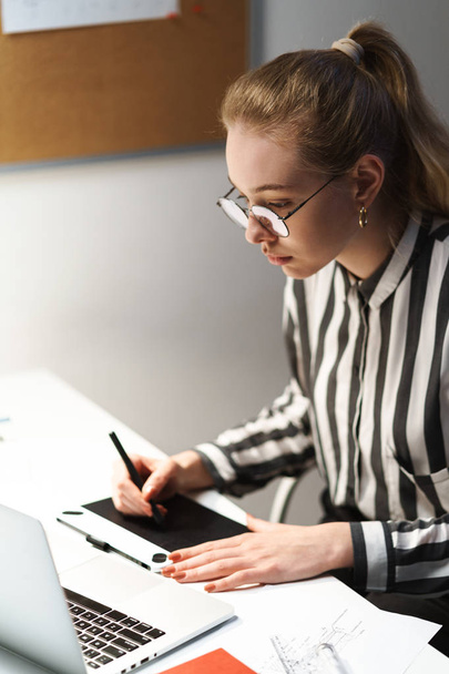 Grafik tablet bilgisayarda taslak tasarımı gözlük giyen ve işyerinde oturan sarışın Avrupalı kadın mimar Fotoğrafı - Fotoğraf, Görsel