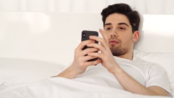 Happy surprised brunette smiing man wearing casual clothes lying in bed and using smartphone - Filmati, video
