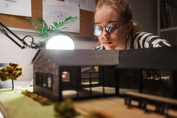 Photo of thoughtful professional woman architect designing draft with house model and sitting at workplace - 写真・画像
