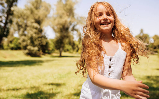 Portrait of happy little girl with long blond hair playing at nature background. Positive child enjoying summer day in the park. Cheerful kid having fun in the forest on sunlight. Happy childhood - 写真・画像