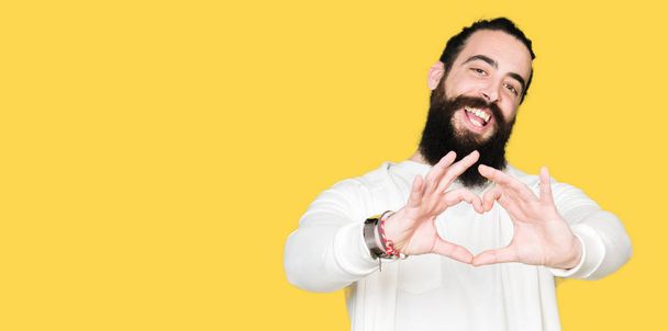 Jonge man met lange haren en baard dragen sportieve sweater glimlachend in liefde met hartsymbool en vorm met de handen. Romantische concept. - Foto, afbeelding