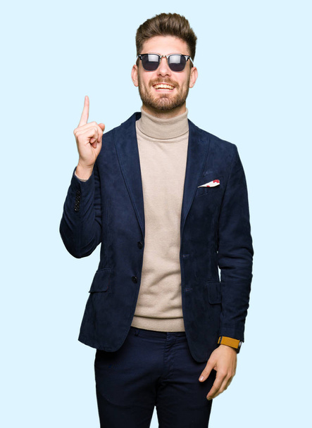 Jonge knappe elegante man dragen van een zonnebril vinger omhoog met succesvolle idee. Verlaten en gelukkig. Nummer één. - Foto, afbeelding