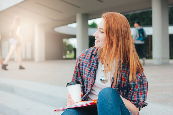 attraktive rothaarige lächelnde Studentin in lässiger Kleidung mit einer Tasse Kaffee in der Hand auf der Straße in der Stadt gehen - Foto, Bild