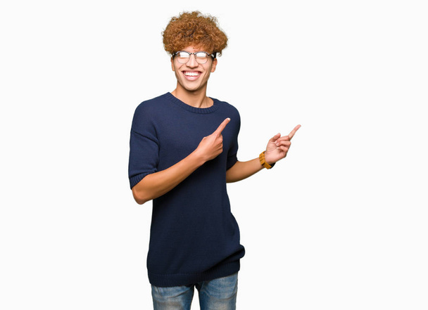 Fiatal jóképű férfi afro szemüveg és mosolyogva néz a kamerába, mutatva a két kéz és az ujjak az oldalon. - Fotó, kép