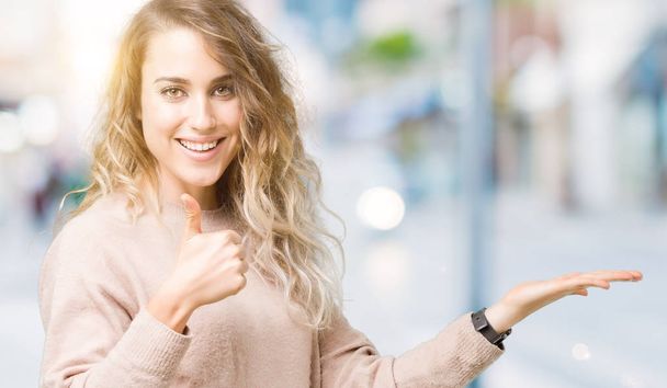 Mooie jonge blonde vrouw sweatershirt over geïsoleerde achtergrond weergegeven: palm hand dragen en doen ok gebaar met duim omhoog, gelukkig en vrolijk lachend - Foto, afbeelding