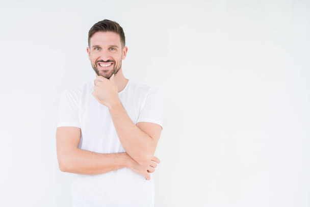 Mladý pohledný muž nosí ležérní bílé tričko izolované pozadí sebevědomým na kameru s úsměvem se zkříženýma rukama a ruku a na bradě. Pozitivní myšlení. - Fotografie, Obrázek