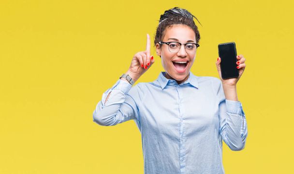 Молодые плетеные волосы африканская бизнес-девушка показывает экран смартфона на изолированном фоне удивлен идеей или вопрос указывая пальцем со счастливым лицом, номер один
 - Фото, изображение