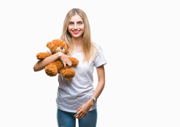 Young beautiful blonde woman holding teddy bear over isolated background with a happy face standing and smiling with a confident smile showing teeth - Foto, immagini