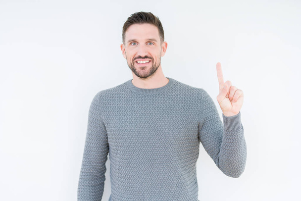 Young handsome man wearing casual sweater over isolated background showing and pointing up with finger number one while smiling confident and happy. - Foto, Imagen