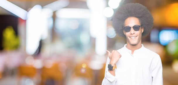 Молодий афроамериканець людиною з афро волосся носити сонцезахисні окуляри, посміхаючись з щасливим обличчям, дивлячись і вказуючи на сторону з великим пальцем. - Фото, зображення