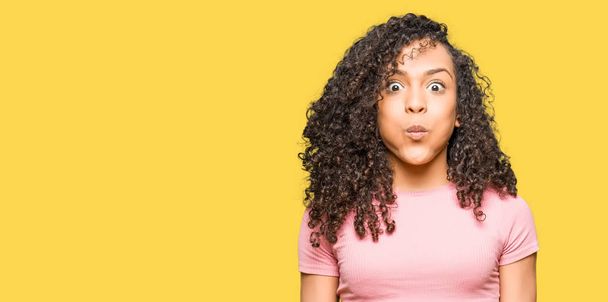 Young beautiful woman with curly hair wearing pink t-shirt puffing cheeks with funny face. Mouth inflated with air, crazy expression. - Photo, Image