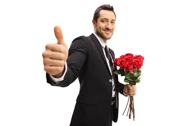 Young elegant man holding a bunch of roses and showing thumbs up isolated on white background - Фото, зображення