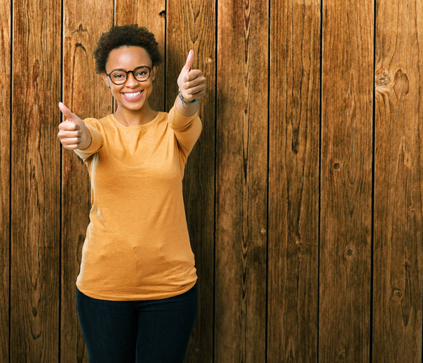 Young beautiful african american woman wearing glasses over isolated background approving doing positive gesture with hand, thumbs up smiling and happy for success. Looking at the camera, winner gesture. - Photo, Image