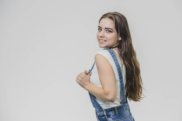 Young pretty student girl standing on a white background. Dressed in a denim suit and a white T-shirt. Holding his hands on the pliers from the combinator eyefully looks into the camera. - Foto, Bild