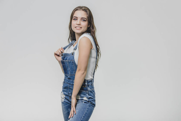 Beautiful girl in a denim overalls and a white T-shirt. Having taken a moment, he put his hand on the placket from the overalls, looking into the camera. Isolated over white background. - Foto, afbeelding