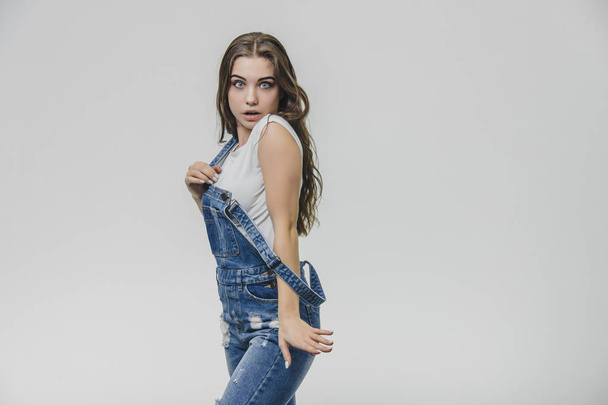 Beautiful girl in a denim overalls and a white T-shirt. Having taken a moment, he put his hand on the placket from the overalls, looking into the camera. - Φωτογραφία, εικόνα