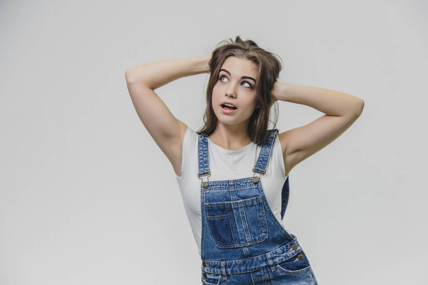 A teenage girl with pleasure an attractive look. Wears fashionable denim overalls and white shirt. Put your hands on his head looking up.Concept of people, style and fashion - Photo, image
