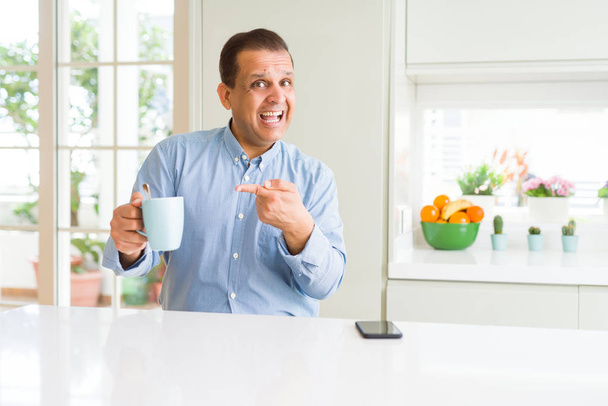 Μεσαίας ηλικίας επιχείρηση άνθρωπος πίνοντας καφέ το πρωί στο σπίτι πολύ χαρούμενος δείχνοντας με το χέρι και το δάχτυλο - Φωτογραφία, εικόνα