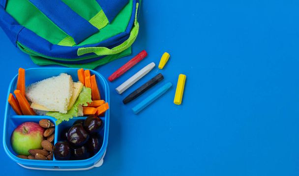 Almuerzo escolar con sándwich, nueces, verduras y frutas sobre fondo azul con útiles escolares. Copiar espacio
. - Foto, Imagen