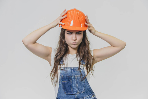 Young pretty girl standing on a white background. During this time wearing a white T-shirt and denim overalls. Dressed in an orange helmet. - Photo, Image
