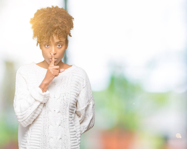 美しい若いアフリカ系アメリカ人女性の唇に指で静かにするように求めて孤立の背景に冬のセーターを着ています。沈黙と秘密の概念. - 写真・画像