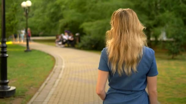 back view. medium plan. beautiful young woman blonde in denim overalls walks through the summer city park - Video, Çekim