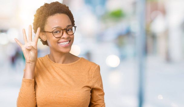 Νέοι όμορφη αφρικανική αμερικανική γυναίκα φοράει γυαλιά πέρα από το απομονωμένο υπόβαθρο δείχνει και δείχνει προς τα επάνω με τα δάχτυλα αριθμός τέσσερα ενώ χαμογελώντας αυτοπεποίθηση και χαρούμενοι. - Φωτογραφία, εικόνα