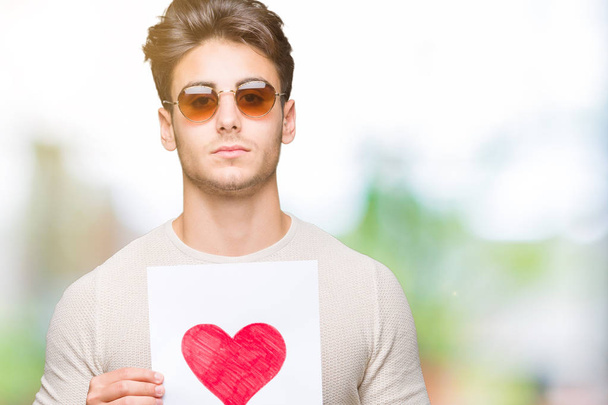 Όμορφος νεαρός κρατώντας κάρτα με κόκκινη καρδιά πάνω από απομονωμένες φόντο με μια αυτοπεποίθηση έκφραση στο έξυπνο πρόσωπο σκέφτεται σοβαρά - Φωτογραφία, εικόνα