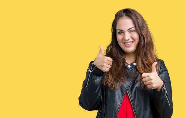 Beautiful plus size young woman wearing a fashion leather jacket over isolated background success sign doing positive gesture with hand, thumbs up smiling and happy. Looking at the camera with cheerful expression, winner gesture. - Fotó, kép