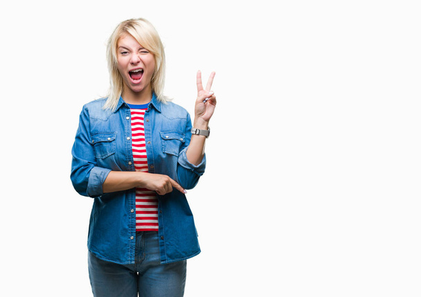 Young beautiful blonde woman wearing denim jacket over isolated background smiling with happy face winking at the camera doing victory sign. Number two. - Fotoğraf, Görsel