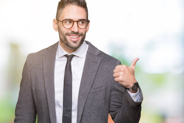 Young handsome business man over isolated background smiling with happy face looking and pointing to the side with thumb up. - Photo, Image
