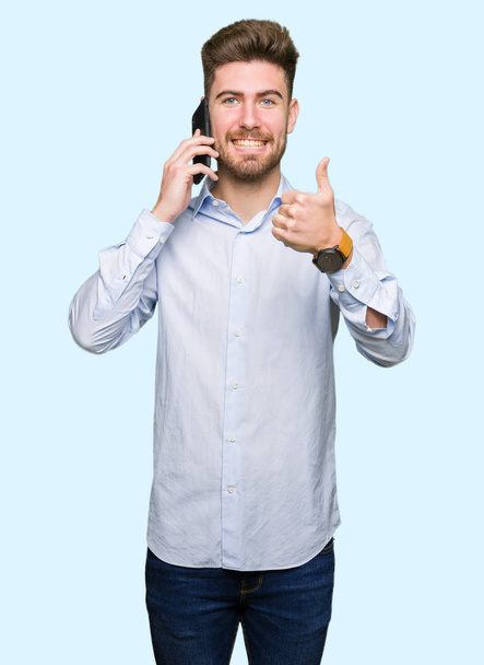 Young handsome man business talking on smartphone happy with big smile doing ok sign, thumb up with fingers, excellent sign - Foto, Imagen