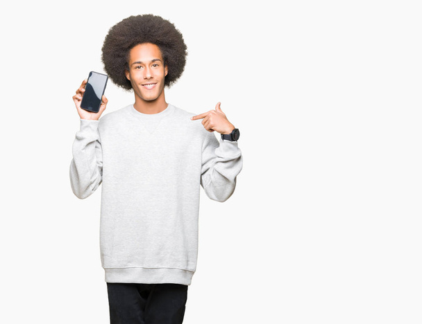 Joven hombre afroamericano con cabello afro mostrando la pantalla del teléfono inteligente con la cara sorpresa apuntando con el dedo hacia sí mismo
 - Foto, Imagen