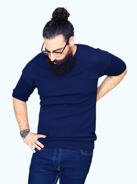 Homem jovem hipster com cabelos longos e barba vestindo óculos Sofrendo de dor nas costas, tocando nas costas com a mão, dor muscular
 - Foto, Imagem