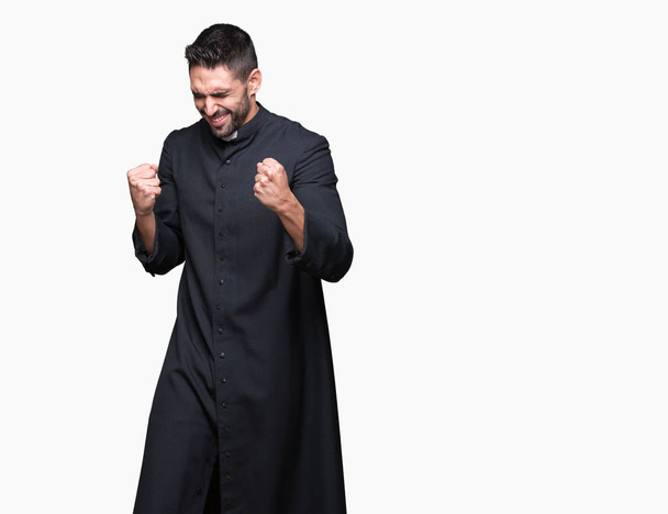 Young Christian priest over isolated background very happy and excited doing winner gesture with arms raised, smiling and screaming for success. Celebration concept. - Photo, Image