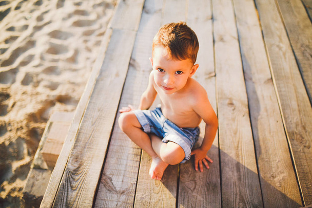 Little Caucasian boy child sitting on wooden pier sandy beach, summer time, sea vacation near water. The theme is the flow of time, a short life, the meaning and purpose of existence. Meaning of life - Photo, Image