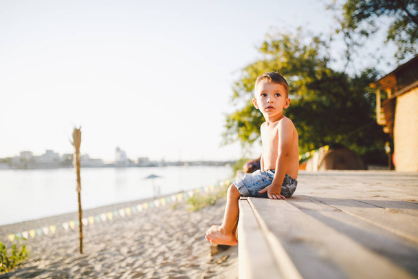 Mały kaukaski chłopiec dziecko siedzi na drewnianej molo piaszczystej plaży, czas letni, morze wakacje w pobliżu wody. Tematem jest przepływ czasu, krótkie życie, sens i cel istnienia. Sens życia - Zdjęcie, obraz