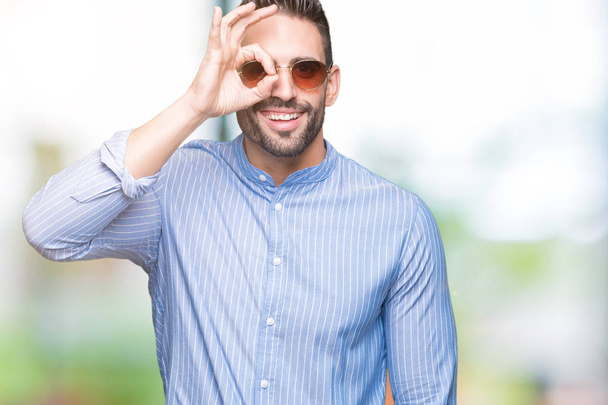 Young handsome man wearing sunglasses over isolated background doing ok gesture with hand smiling, eye looking through fingers with happy face. - Photo, Image